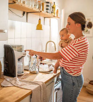 Eine Frau bereitet in der Küche Nahrung mit einem elektrischen Babykostwärmer zu, während sie ihr Kind auf dem Arm hält.