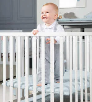 Ein lachendes Baby steht sicher in einem Holz-Laufgitter und hält sich an den Stäben fest.