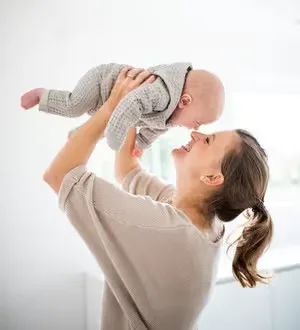 Eine lachende Mutter hebt ihr Baby, das einen gemütlichen Strickstrampler trägt.