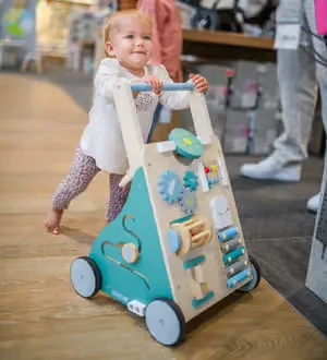 Ein Kleinkind schiebt einen bunt gestalteten Lauflernwagen aus Holz, der mit verschiedenen motorischen Spielelementen ausgestattet ist.
