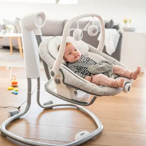 Ein Baby liegt entspannt in einer elektrischen Babywippe mit Spielbogen in einem Wohnzimmer.