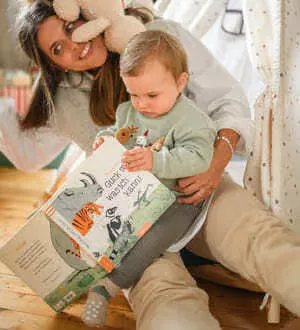 Eine Mutter und ein Kleinkind sitzen gemütlich am Boden und schauen sich gemeinsam ein großes, illustriertes Buch an.