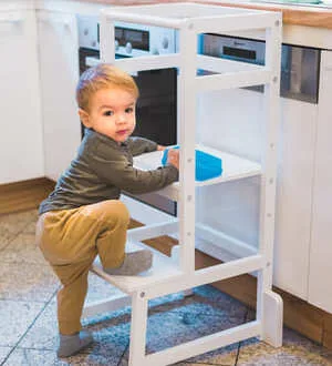 Ein kleiner Junge klettert in der Küche auf einen hölzernen Lernturm, um sicher die Arbeitsplatte zu erreichen.