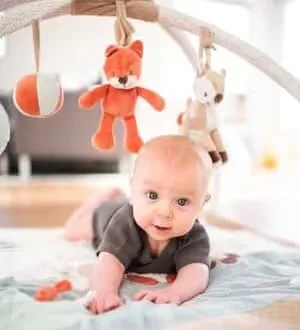 Ein Baby liegt auf einer Spielmatte unter einem Spielbogen mit hängenden Stofftieren wie einem Fuchs und einem Reh.
