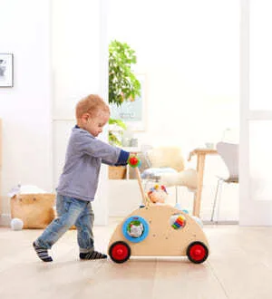 Ein kleiner Junge schiebt einen hölzernen Lauflernwagen mit bunten Rädern und integrierten Spielelementen durch ein helles Zimmer.
