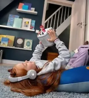 Ein Kind liegt mit Kopfhörern auf dem Boden und stellt eine Tonie-Figur auf eine Toniebox, um ein Hörspiel zu starten.