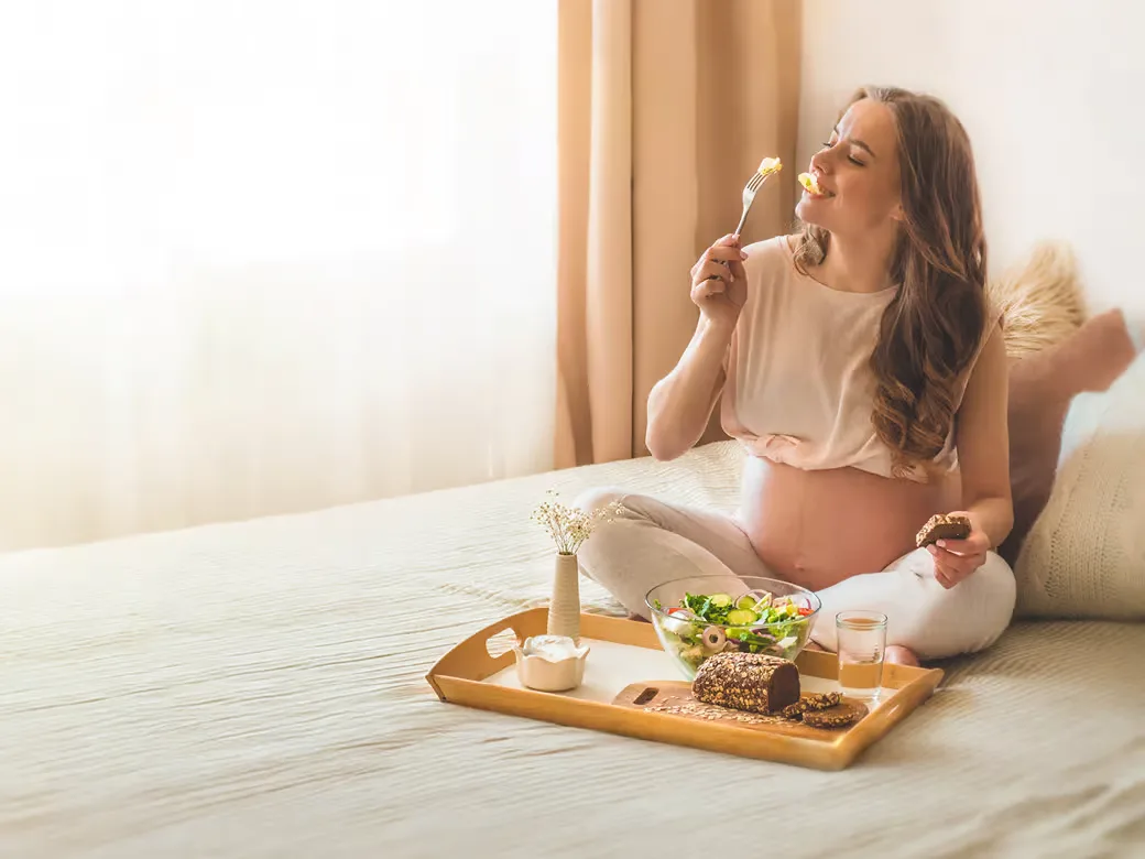 Schwangere Frau sitzt auf dem Bett, hält eine Gabel in der Hand und isst. Vor ihr steht ein Tablett mit Essen.