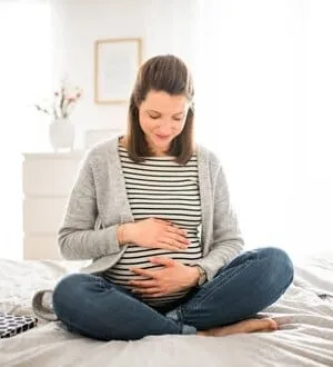 Eine schwangere Frau sitzt mit einem bequemen Outfit auf einem Bett und hält lächelnd ihre Hände auf ihren Babybauch.